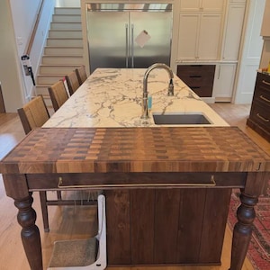 May include: A rectangular wooden kitchen island featuring a thick end-grain butcher block top with a multi-toned checkerboard pattern. The base is constructed from dark wood with turned legs, a horizontal metal handle, and the letter J engraved on the side.