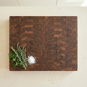 May include: A rectangular end-grain butcher block cutting board with a dark brown wood pattern. Fresh herbs and a pile of salt are arranged on the board. The bottom right corner has the word "cey" printed on it.