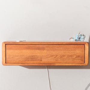 May include: A wall-mounted wooden shelf with a drawer, made from light brown wood. The shelf has a rounded edge. A small, colourful glass sculpture and a white cord are on the shelf.