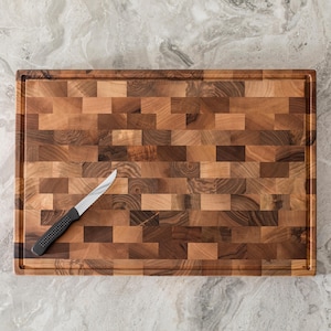 May include: Rectangular wooden cutting board with a mosaic pattern of various wood tones. A small serrated knife with a black handle rests on the board. The board has a juice groove around the perimeter.