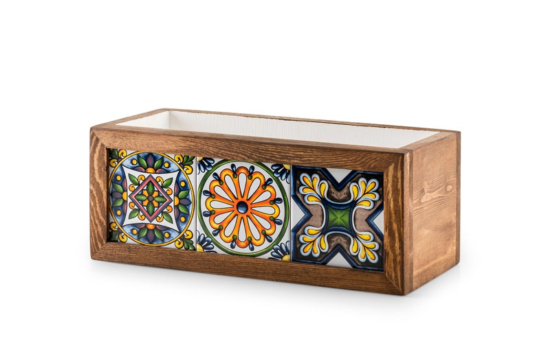 May include: Rectangular wooden planter box with a dark brown finish. The front features three decorative ceramic tiles with colorful floral and geometric patterns. The interior is lined with white material.