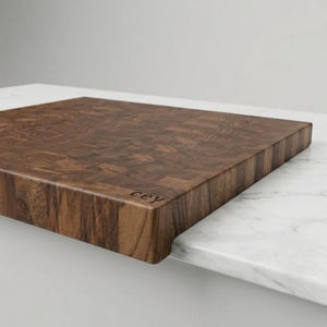 May include: A dark brown end-grain chopping board with a textured surface. The board has a smooth edge and the letters "cey" are engraved on the corner. It is placed on a white surface. The board is likely made of hardwood.