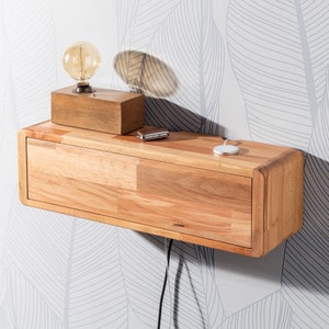 May include: A floating wooden nightstand with a drawer, a wooden lamp, and a wireless charger. The nightstand is made of light-colored wood and is mounted on a wall with a leaf pattern wallpaper. A light bulb is in the lamp.