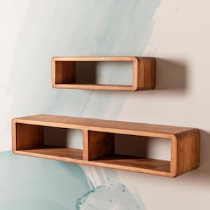 Può includere: Due mensole galleggianti in legno con finitura marrone caldo sono montate su un muro. La mensola superiore è un'unica scatola rettangolare, mentre la mensola inferiore ha due scomparti. Le mensole hanno bordi arrotondati.