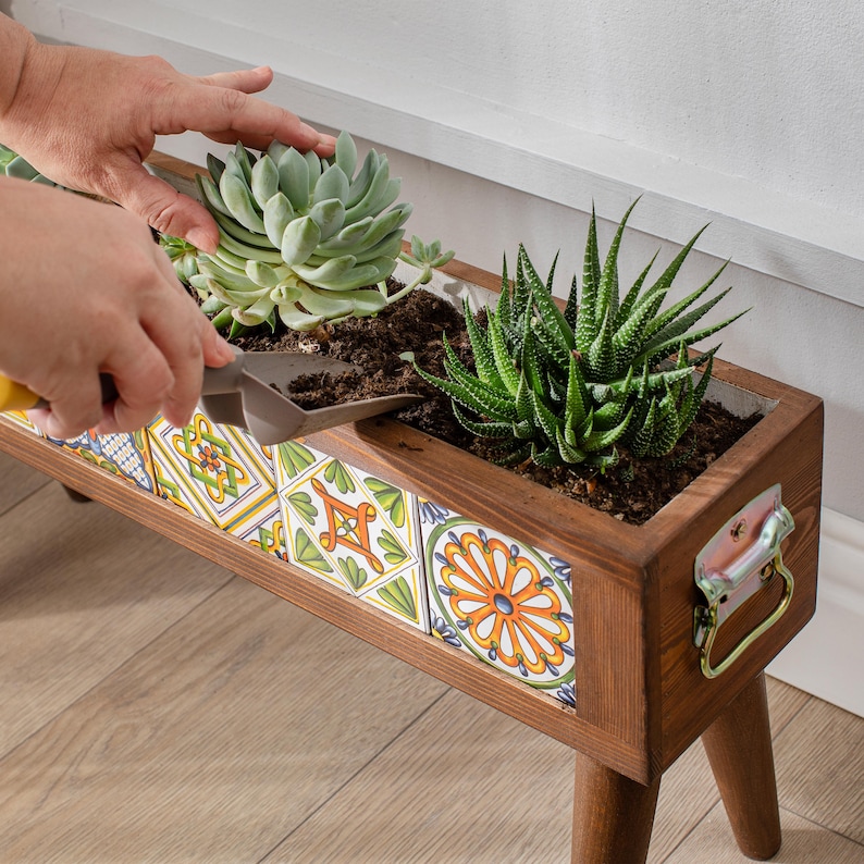 May include: A wooden planter box with a colorful tile design. The planter is filled with soil and succulents. The planter has four legs and a metal handle.