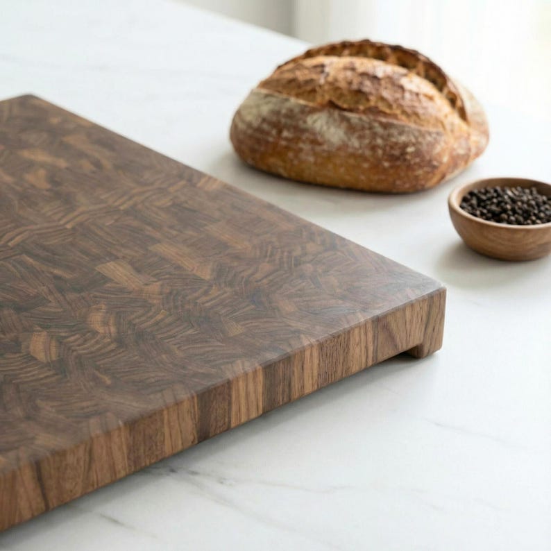 May include: A dark brown wooden cutting board with a textured surface. A loaf of bread and a small wooden bowl of peppercorns are in the background. The cutting board has a rectangular shape and a raised edge.