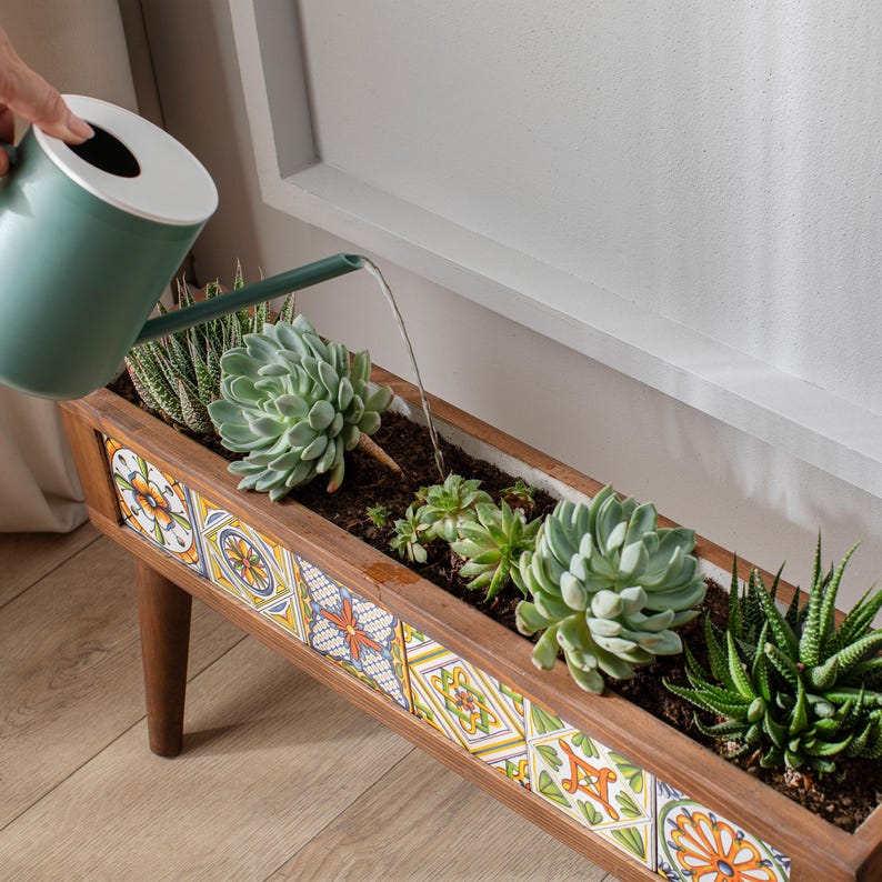 May include: A wooden planter box with colorful tile accents is filled with succulents and being watered with a green watering can. The planter has a rustic wooden stand.