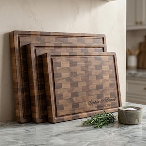 May include: Three wooden end-grain chopping boards in different sizes, featuring a dark brown and tan chequered pattern. The largest board has a carved border and the names "Sophia & Daniel" with the date "October 15, 2024" engraved. A sprig of rosemary and a small ceramic bowl are in the foreground.