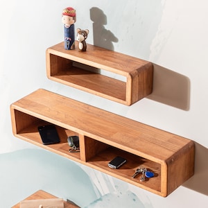 May include: Two wooden floating shelves with rounded edges are mounted on a wall. The top shelf holds a small figurine and a bear-shaped ornament. The lower shelf contains a smartphone, keys, and a car key fob. The shelves are made of light brown wood.