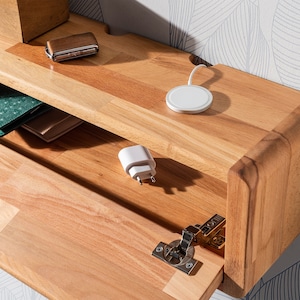 May include: A wooden shelf with a hidden compartment, featuring a wireless charging pad, a phone charger, and a wallet. The shelf is made of light wood with a natural finish.