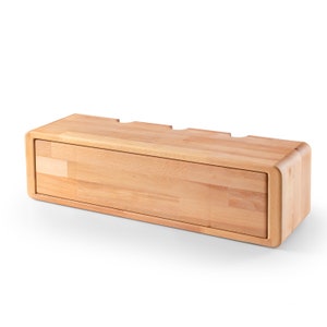 May include: A light wood wall-mounted shelf with a hinged door and three cutouts on the top. The shelf is made of natural wood with a light finish.
