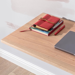 Pu&ograve; includere: Una scrivania in legno chiaro con un diario rilegato in pelle rossa, un libro pi&ugrave; piccolo con una copertina colorata e una penna rossa in cima. Un laptop argentato &egrave; sulla scrivania.
