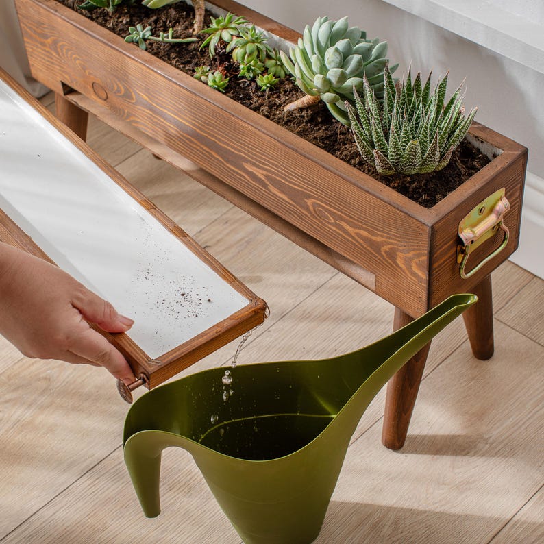 May include: A wooden planter box with a white liner and a watering can pouring water into the planter. The planter is on a stand with legs and has a brass handle. The planter is filled with succulents and other plants.