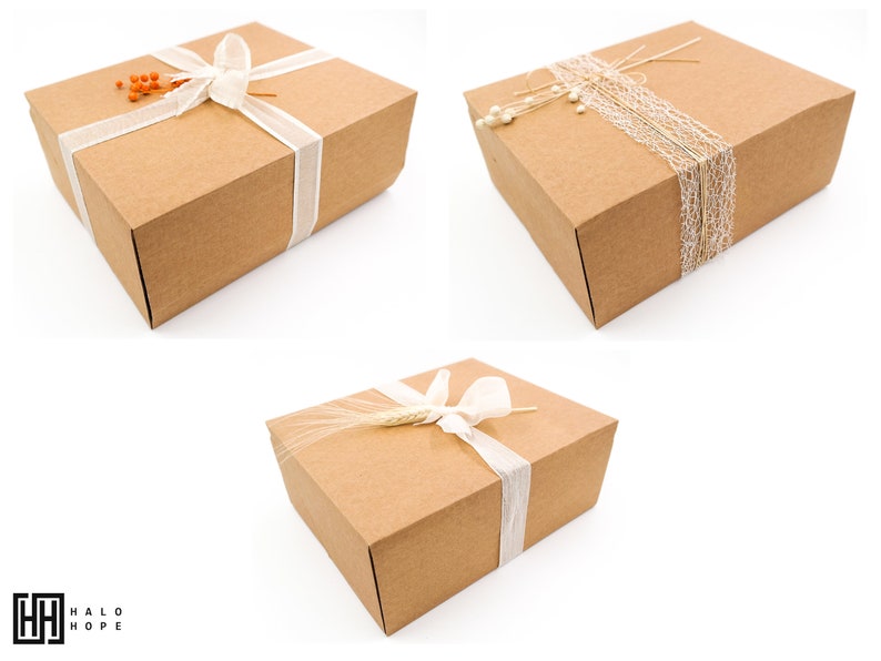 Puede incluir: Tres cajas de regalo de cart&oacute;n marr&oacute;n con diferentes elementos decorativos. La primera caja est&aacute; atada con una cinta blanca y decorada con bayas naranjas. La segunda caja est&aacute; decorada con encaje blanco y cordel. La tercera caja est&aacute; decorada con una cinta blanca y una espiga de trigo.