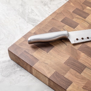 May include: A stainless steel kitchen knife with a wooden handle sits on a brown wooden chopping board with a checkered pattern.