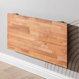 May include: A rectangular, wall-mounted folding table made of light brown wood. The table is attached to the wall with black metal hinges. The wood grain is visible, and the table is mounted above a white baseboard.