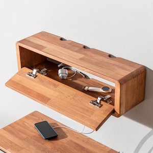 May include: A wooden wall-mounted charging station with a hinged door. Inside, charging cables and a smartwatch are visible. A smartphone rests on a matching wooden surface below, connected by a charging cable. The unit is made of wood.