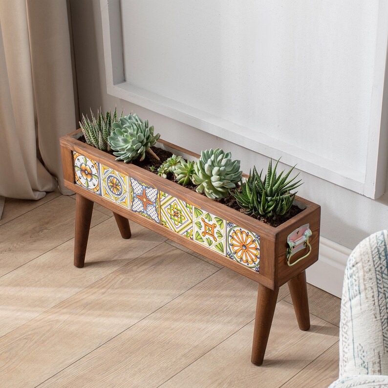May include: A wooden planter with a colorful tile design and four legs. The planter is filled with succulents and is sitting on a wooden floor.