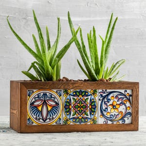 May include: A rectangular wooden planter box with three colorful, patterned ceramic tiles. The box contains green aloe vera plants. The tiles feature intricate designs in blue, yellow, and red. The wood has a warm, brown tone.