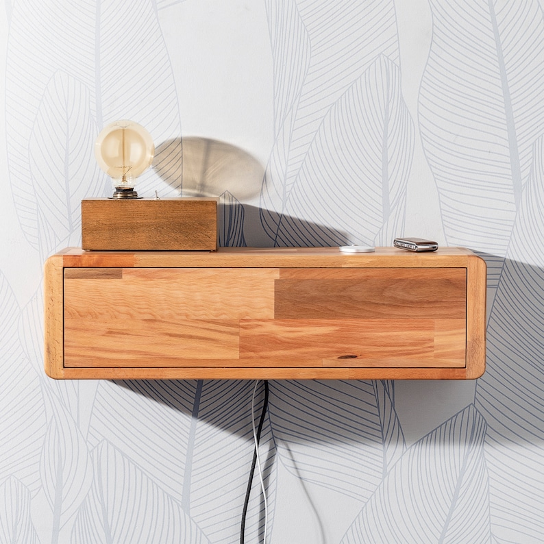 May include: A light wood floating shelf with a drawer, a small lamp, and a phone on top. The shelf is mounted on a white wall with a light blue and white patterned wallpaper.