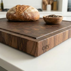 May include: A dark wood cutting board with a loaf of bread and a small wooden bowl of peppercorns. The board has a rectangular shape with a carved edge and the engraved letters "cey" on the side. The bread is round with a golden-brown crust.