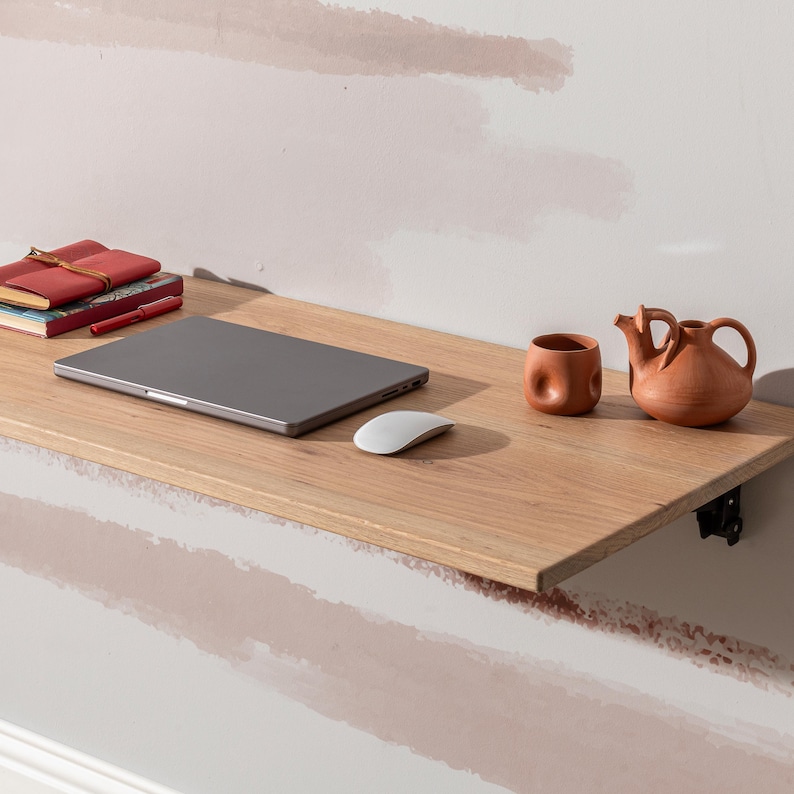 Pu&ograve; includere: Una scrivania pieghevole in legno chiaro con un laptop, un mouse wireless, una penna rossa, una pila di quaderni e due pezzi di ceramica in terracotta in cima.