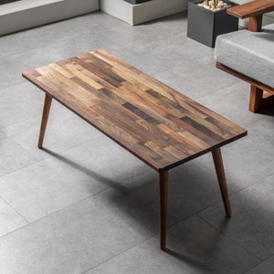 Peut inclure: Une table basse rectangulaire en bois avec un fini grain de bois naturel. Le plateau est composé de diverses pièces de bois, créant un motif unique. Elle possède quatre pieds effilés et inclinés. La table mesure environ 122 cm de long.