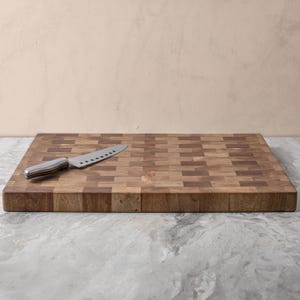 May include: A large, rectangular wooden chopping board with a chequered pattern. The board is made of light and dark wood, creating a contrasting effect. A silver knife is placed on the board.