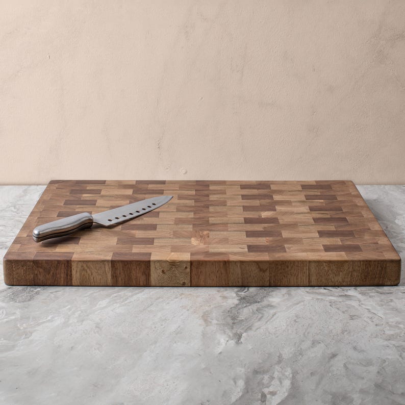 May include: A large, rectangular wooden chopping board with a chequered pattern. The board is made of light and dark wood, creating a contrasting effect. A silver knife is placed on the board.