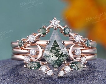 Può includere: Un anello in oro rosa con una pietra di agata verde muschio, circondata da diamanti più piccoli e pietre verdi. L'anello è su uno sfondo grigio.