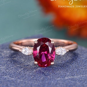 May include: A rose gold engagement ring with a large oval ruby gemstone and two smaller pear-shaped diamonds on either side.