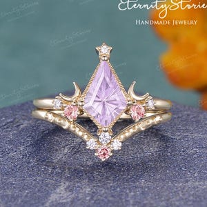 May include: A gold-toned ring set featuring a light purple kite-shaped gemstone, accented with small pink and white gemstones. The ring has a unique design with crescent moon details and a star at the top. The words "Eternity Stories Handmade Jewelry" are in the image.