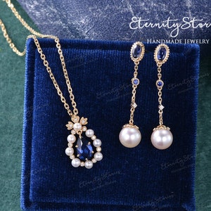 Op de afbeelding: Een gouden ketting met een peer-vormige blauwe saffier hanger omringd door witte parels. De ketting wordt getoond in een blauwe fluwelen doos. Een bijpassend paar oorbellen met blauwe saffieren en witte parels worden ook getoond.