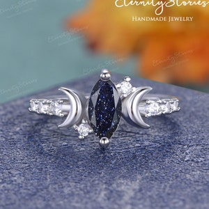 Könnte beinhalten: Ein silberner Ring mit einem blauen Edelstein und zwei Halbmond-Akzenten. Der Edelstein ist im Marquise-Schliff gehalten und hat ein funkelndes, sternenartiges Aussehen. Der Ring ist mit kleinen, klaren Edelsteinen um die Band herum besetzt.