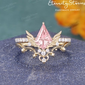 May include: A gold engagement ring with a pink gemstone center and a diamond-studded band. The ring features a unique design with wing-like accents and additional small diamonds. The words "Eternity Stories Handmade Jewelry" are in the background.