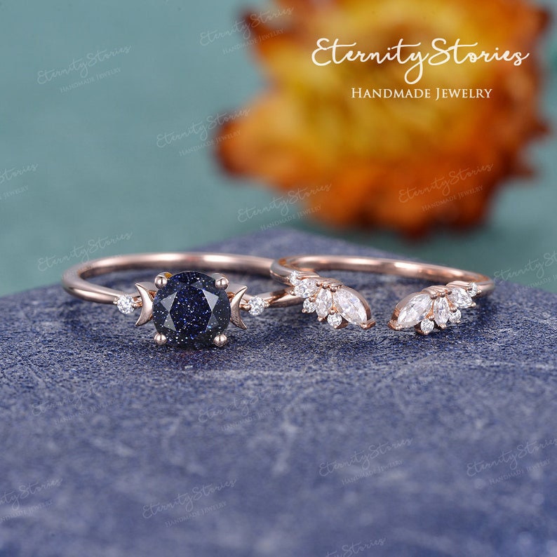 May include: A rose gold ring set with a blue starry sky gemstone and a matching rose gold band with a crescent moon and small diamonds. The band is designed to fit snugly around the engagement ring.