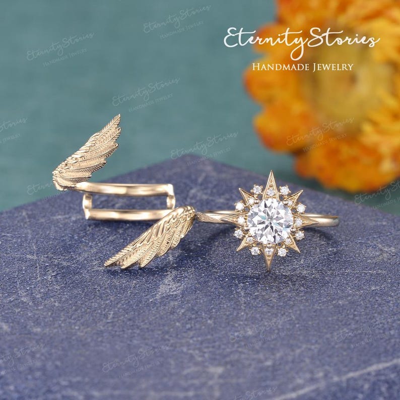 Puede incluir: Un anillo dorado con un dise&ntilde;o de estrella, con una piedra transparente central rodeada de piedras m&aacute;s peque&ntilde;as. Junto al anillo hay un anillo dorado en forma de ala. El fondo es una superficie azul oscuro con el texto "Eternity Stories Handmade Jewelry".