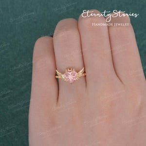 Pu&ograve; includere: Un anello color oro con una gemma rosa a forma di cuore con un disegno a stella. L'anello presenta dettagli di ali e mezzaluna. Le parole "Eternity Stories" e "HANDMADE JEWELRY" sono visibili sullo sfondo.