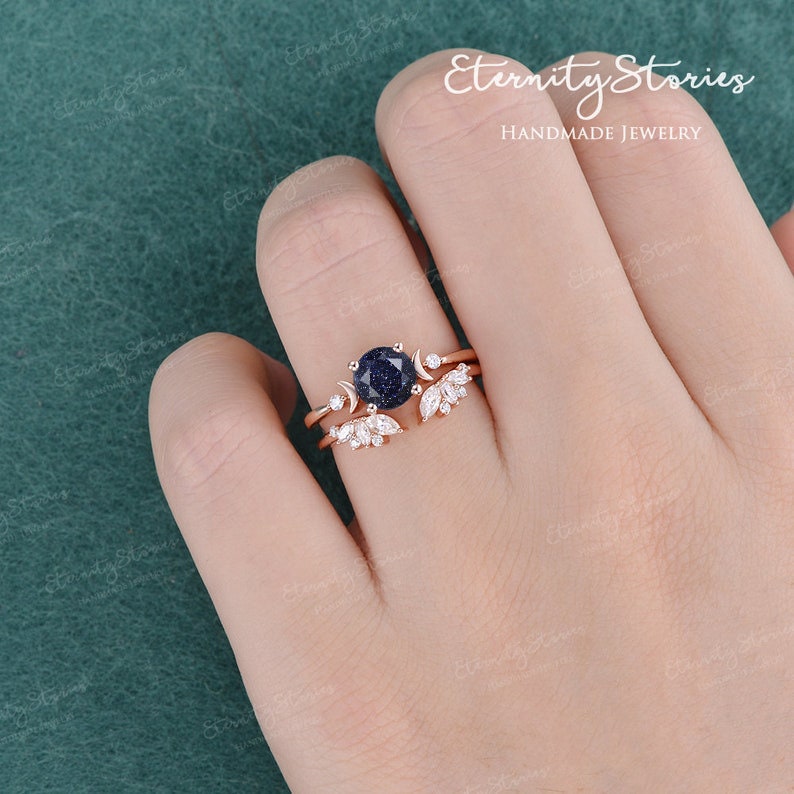 May include: A rose gold ring set with a blue gemstone and accented with small white gemstones. The ring features a crescent moon design and delicate leaf details.  Eternity Stories Handmade Jewellery.