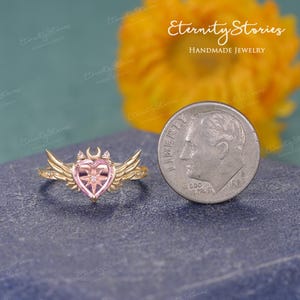 Pu&ograve; includere: Un anello color oro con una pietra centrale rosa a forma di cuore, una mezzaluna e dettagli a stella. L'anello presenta dettagli a forma di ali su entrambi i lati. Una moneta &egrave; accanto all'anello per il confronto delle dimensioni.