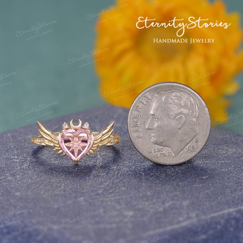 Pu&ograve; includere: Un anello color oro con una pietra centrale rosa a forma di cuore, una mezzaluna e dettagli a stella. L'anello presenta dettagli a forma di ali su entrambi i lati. Una moneta &egrave; accanto all'anello per il confronto delle dimensioni.