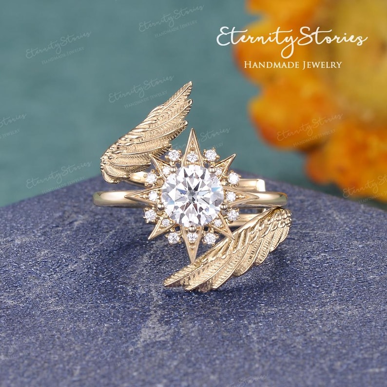 Puede incluir: Un anillo dorado con una piedra central redonda y transparente grande, rodeada de piedras m&aacute;s peque&ntilde;as. El anillo presenta un dise&ntilde;o de estrella y detalles en forma de pluma. El texto "Eternity Stories Handmade Jewelry" es visible.