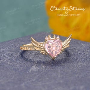 Pu&ograve; includere: Un anello color oro con una gemma rosa a forma di cuore, ornato da ali e una mezzaluna. L'anello presenta un design a stella all'interno del cuore e piccole pietre di accento. Il testo "Eternity Stories Handmade Jewelry" &egrave; visibile.