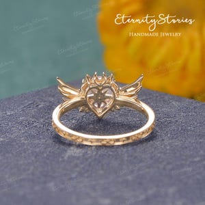 Pu&ograve; includere: Un anello color oro con una pietra centrale a forma di cuore, fiancheggiata da ali e un design a corona. La fascia dell'anello presenta un motivo inciso. Lo sfondo include il testo "Eternity Stories Handmade Jewelry".