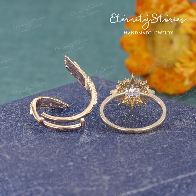 Puede incluir: Dos anillos dorados sobre una superficie azul. Uno presenta un dise&ntilde;o de estrella con una piedra central, mientras que el otro tiene un elemento curvo en forma de pluma. El texto "Eternity Stories Handmade Jewelry" es visible.