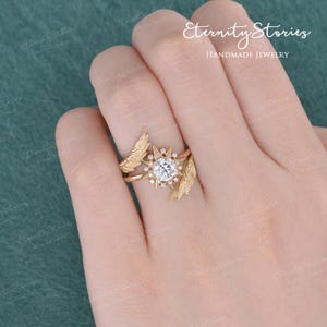 Puede incluir: Un juego de anillos dorados con un anillo central con una gema grande, redonda y transparente rodeada de piedras m&aacute;s peque&ntilde;as en un dise&ntilde;o de estrella. El juego incluye una banda con detalles en forma de pluma. El texto "Eternity Stories Handmade Jewelry" es visible.