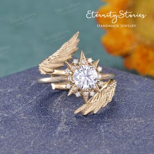 Puede incluir: Un anillo dorado con una gema grande, redonda y transparente en el centro, rodeada de piedras transparentes m&aacute;s peque&ntilde;as. El anillo presenta un dise&ntilde;o de estrella y detalles en forma de pluma. El texto "Eternity Stories Handmade Jewelry" es visible.
