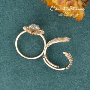 Puede incluir: Juego de anillos de oro de dos piezas. Un anillo presenta una piedra preciosa transparente central rodeada de un dise&ntilde;o floral. El segundo anillo tiene un dise&ntilde;o curvo y texturizado. Los anillos se muestran sobre una superficie turquesa. El texto "Eternity Stories Handmade Jewelry" es visible.