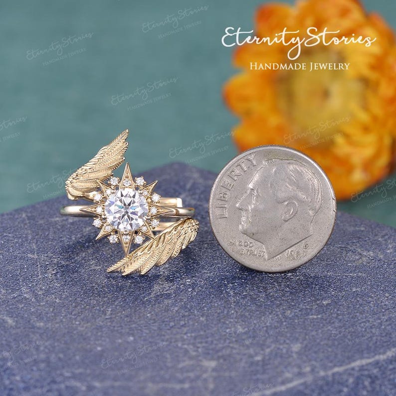 Puede incluir: Un anillo dorado con una gema central redonda y transparente, rodeada de piedras m&aacute;s peque&ntilde;as y un dise&ntilde;o de estrella. El anillo presenta detalles en forma de plumas a ambos lados. El texto "Eternity Stories HANDMADE JEWELRY" es visible.