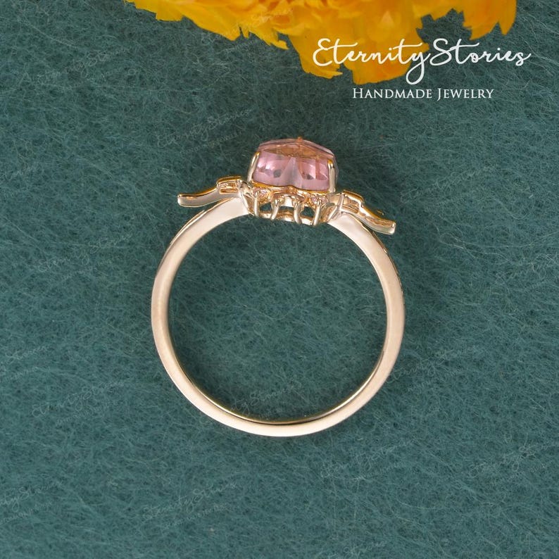 Pu&ograve; includere: Un anello d'oro con una pietra rosa al centro e dettagli decorativi. L'anello &egrave; posto su uno sfondo verde con il testo "Eternity Stories Handmade Jewelry".